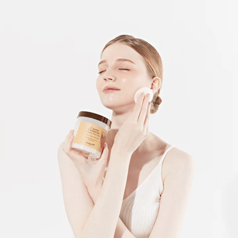Una donna con i capelli raccolti applica un prodotto per la cura della pelle sul viso con un batuffolo di cotone. Tiene un grande barattolo etichettato "Carrot Carotene Calming Water Pad". I suoi occhi chiusi e l'espressione pacifica trasmettono una sensazione di relax e piacere nella routine di cura della pelle.