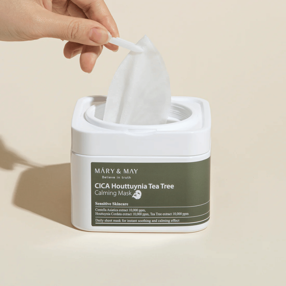 Mano che tira su una maschera in tessuto dal barattolo Cica Houttuynia Tea Tree Calming Mask di Mary & May – lenitiva maschera viso per pelle sensibile con Cica e estratto di Tea Tree.