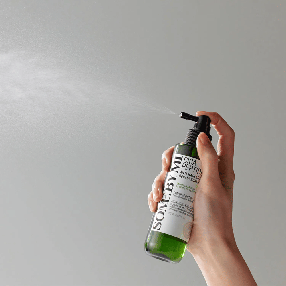 Mano che spruzza SOME BY MI Cica Peptide Anti Hair Loss Derma Scalp Tonic da una bottiglia verde con ugello nero ed etichetta bianca su sfondo grigio.