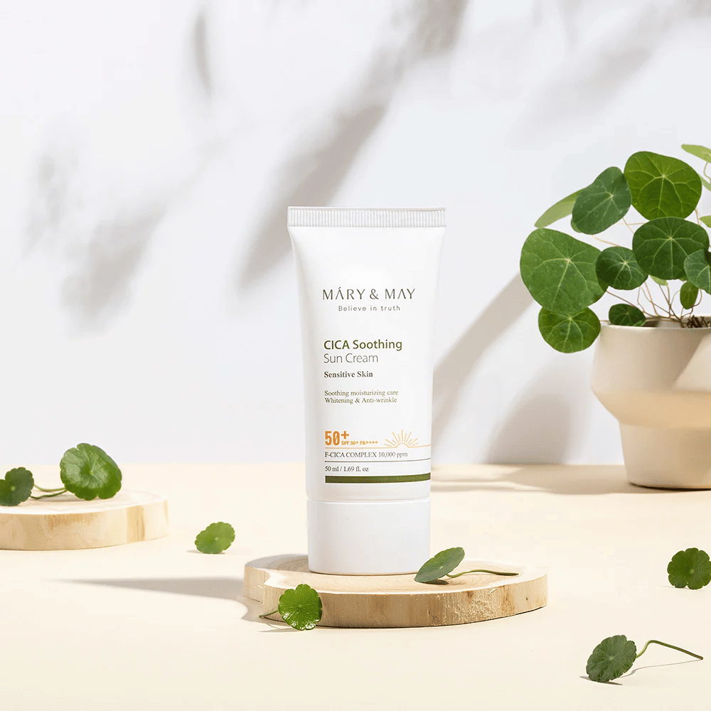 Mary&May CICA Soothing Sun Cream posizionata su un podio in legno con foglie verdi di centella tutt'intorno. L'immagine enfatizza gli ingredienti lenitivi per la pelle e l'ispirazione naturale alla base del prodotto.