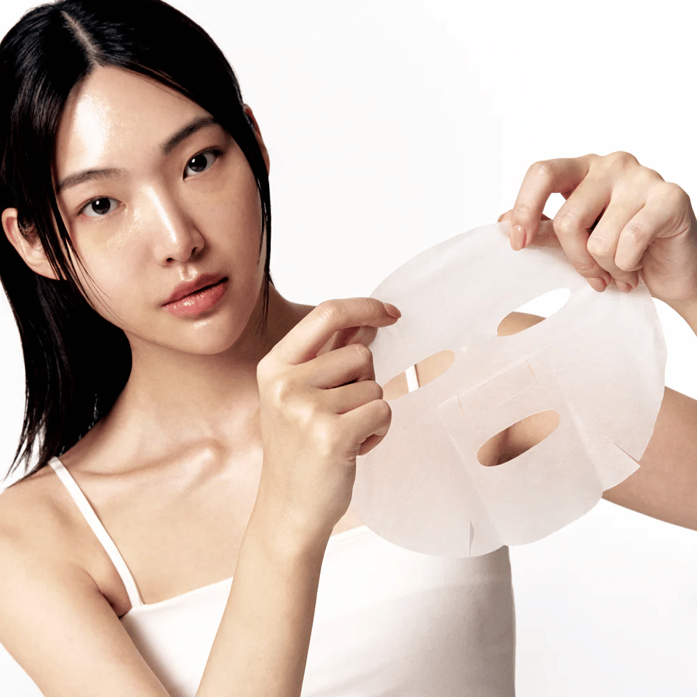 Donna con pelle pulita e idratata tiene un sheet mask bianco di fronte a sé, pronta ad applicarlo sul viso.