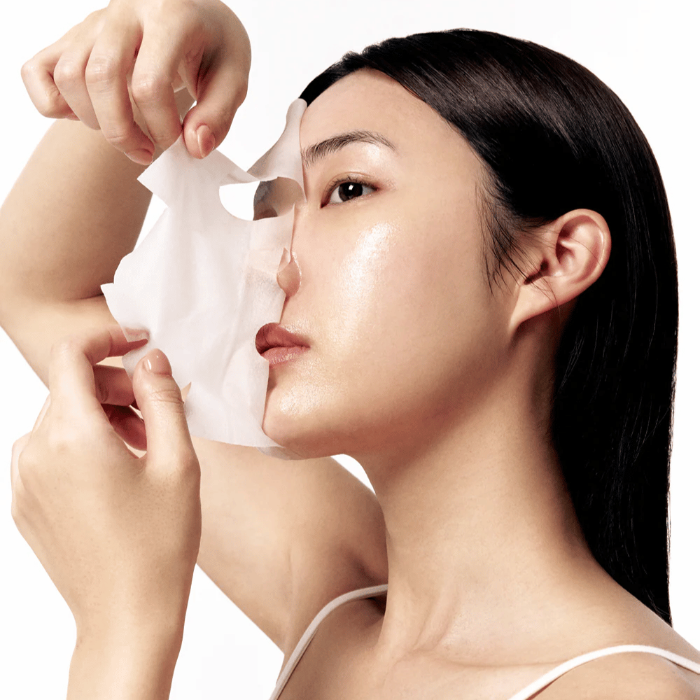 Donna con pelle liscia e idratata rimuove un sheet mask bianco dal viso, rivelando un incarnato radioso e uniforme.