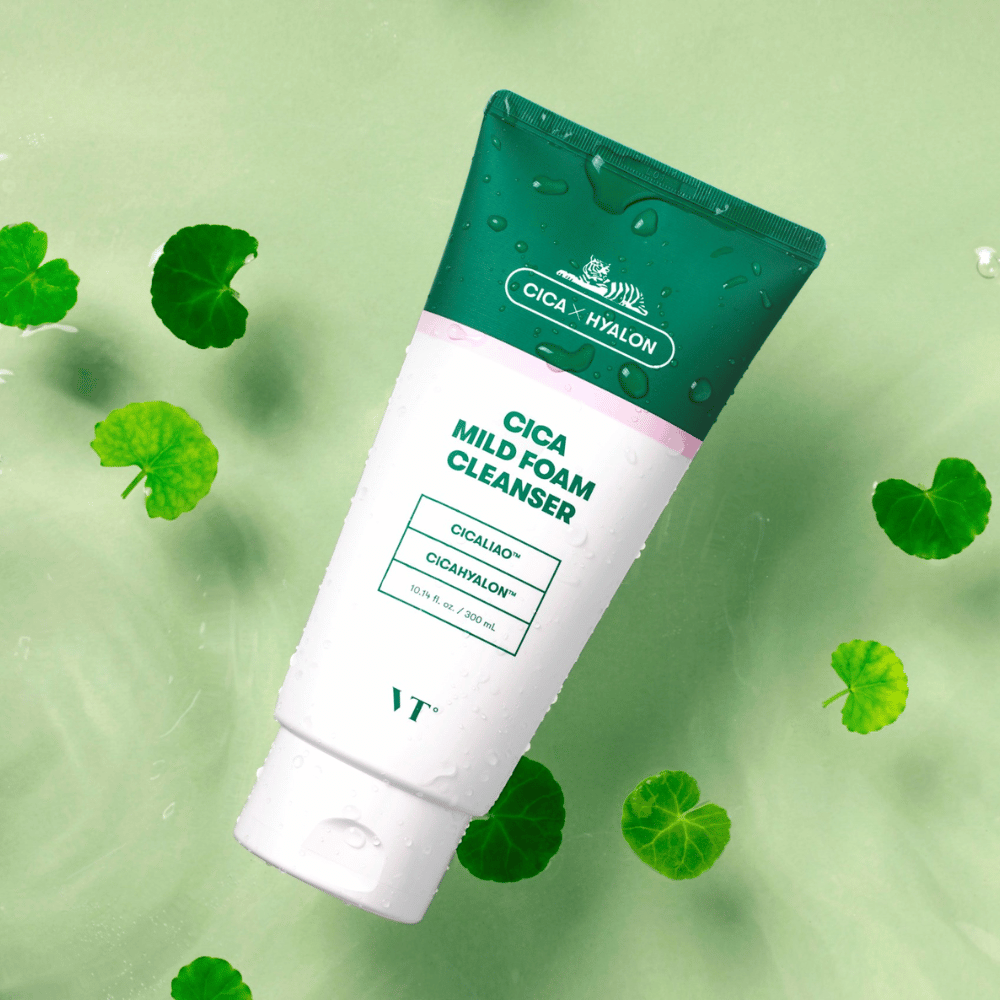 VT Cica Mild Foam Cleanser posizionato su una superficie verde chiaro con gocce d'acqua sulla confezione e circondato da foglie di centella asiatica verde, il che evidenzia le proprietà calmanti del prodotto.