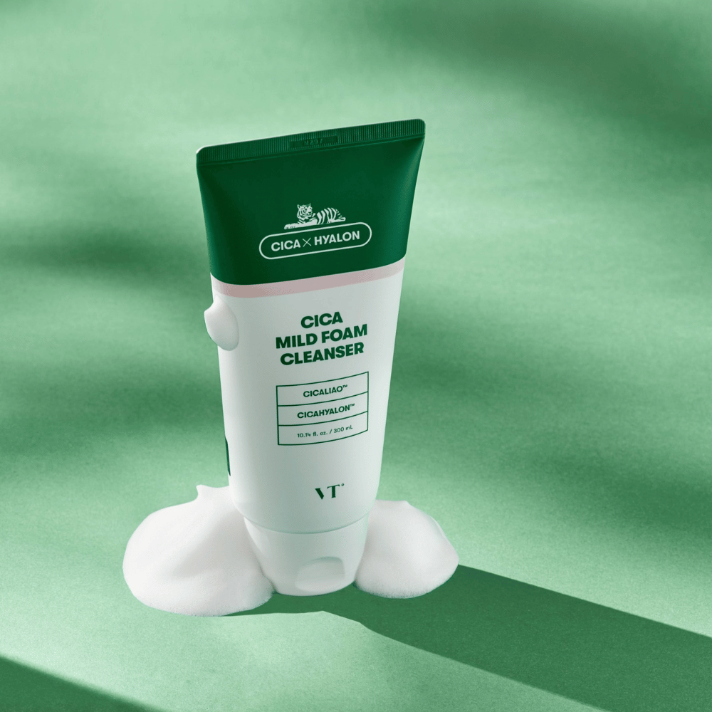 Tubo con VT Cica Mild Foam Cleanser in piedi su una superficie verde con schiuma intorno alla base. Il prodotto ha un design verde e bianco ed è commercializzato con CICALIAO™ e CICAHYALON™.