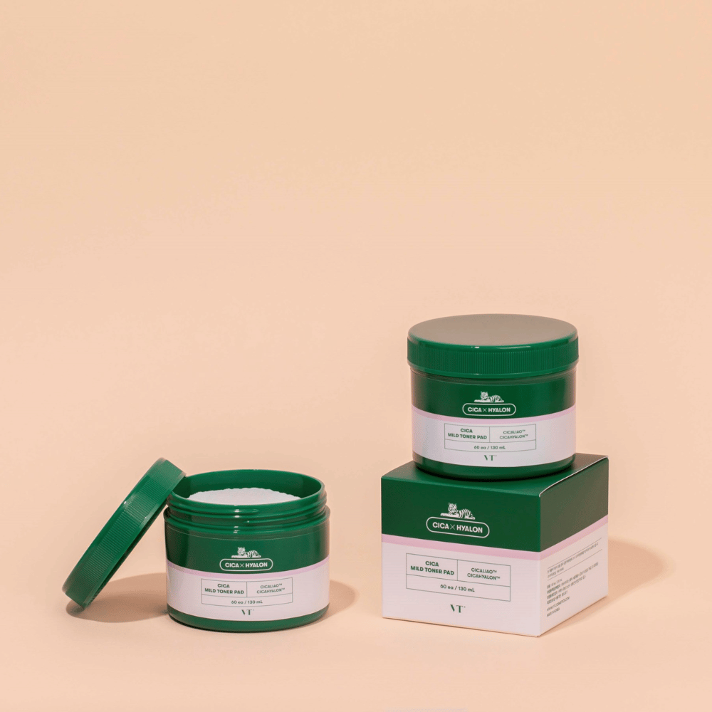 Due barattoli verdi con VT Cica Mild Toner Pad, uno aperto che mostra i dischetti bianchi e uno chiuso posizionato sopra la sua confezione, su sfondo beige.