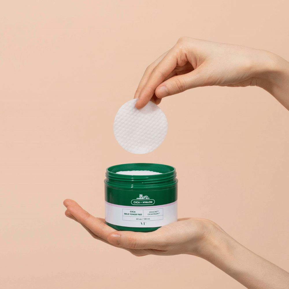 Mano che tiene un barattolo verde con VT Cica Mild Toner Pad e solleva un dischetto bianco rotondo sopra il barattolo su sfondo beige.