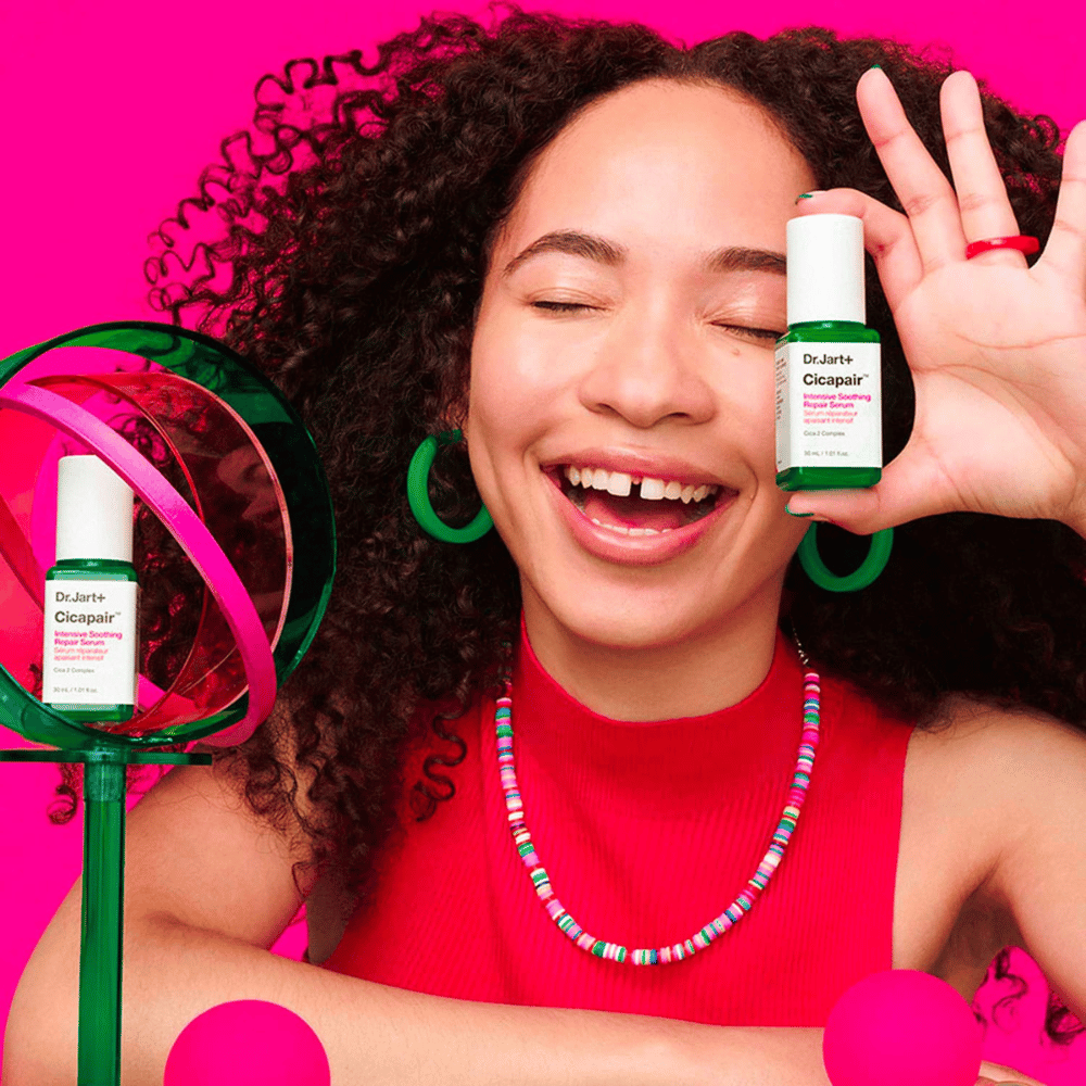 Una donna sorride ampiamente mentre tiene Dr.Jart+ Cicapair Intensive Soothing Repair Serum davanti al suo viso. Indossa orecchini verdi e una collana rossa. Lo sfondo è rosa chiaro e decorato con cerchi verdi e rosa.