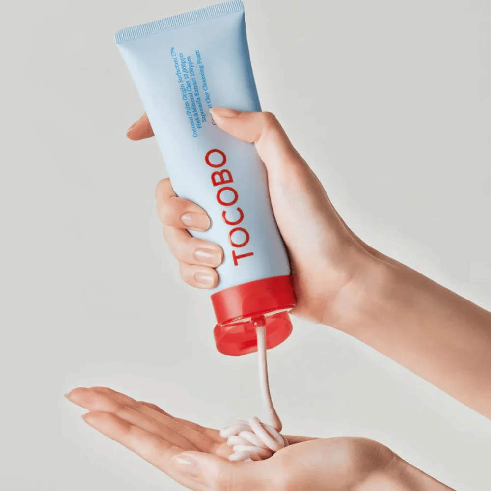 Una persona tiene in mano un tubo di TOCOBO Coconut Clay Cleansing Foam. Il tubo è azzurro chiaro con un tappo rosso e la scritta "TOCOBO" sul davanti. Il prodotto viene spremuto dal tubo e si forma come uno shampoo nella mano. Lo sfondo è neutro e mette a fuoco il prodotto.