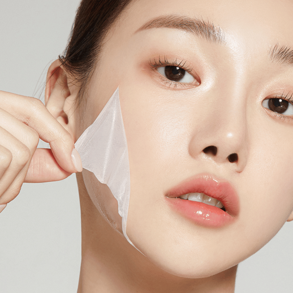 L'immagine mostra una giovane donna che rimuove una sottile maschera viso, mettendo in risalto la sua pelle idratata e morbida. La maschera segue i contorni del viso, dando l'impressione di un prodotto per la cura della pelle efficace e delicato. La sua espressione è rilassata.