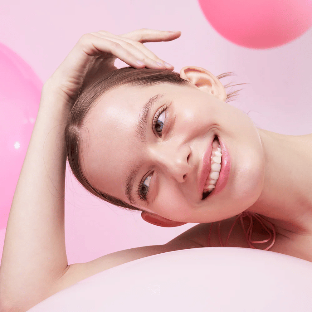 En kvinna ler och vilar sitt huvud på handen, omgiven av rosa luftballonger. Hennes hud ser strålande och naturligt frisk ut.
