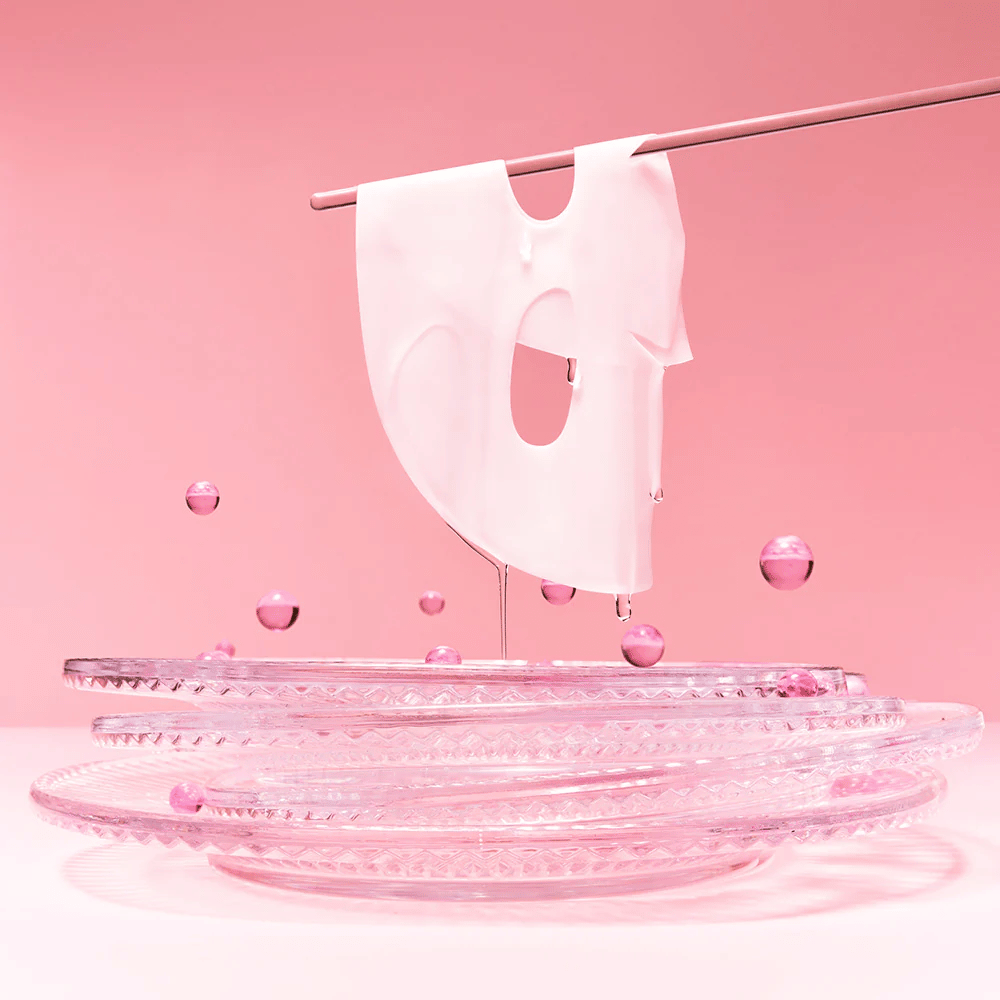 En hydrogelmask hänger på en stång, droppande med serum. Bakgrunden är rosa och spegeln på botten reflekterar de rosa bubblorna som omger masken.