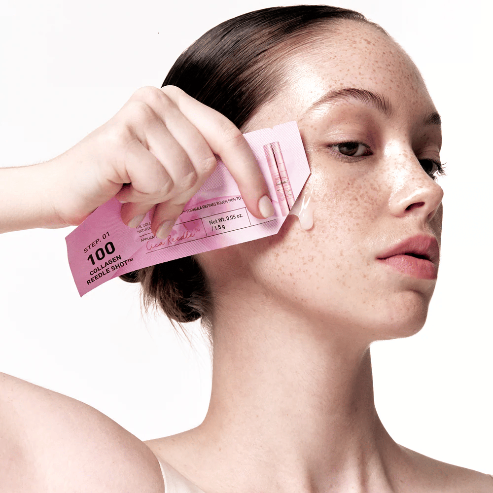 Donna applica il passo 1 di Collagen Reedle Shot 100 con una confezione rosa monouso sulla guancia. Un gel trasparente si vede sulla pelle, segnando il primo passo nel trattamento skincare a due fasi.