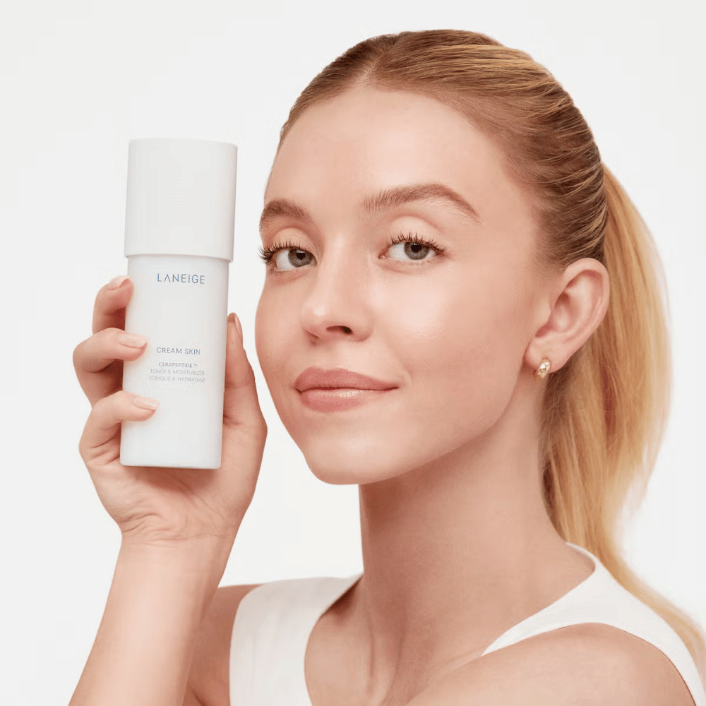 Una persona tiene in mano Laneige Cream Skin Cerapeptide Refiner in una bottiglia bianca. La persona sorride e mostra il prodotto, che è commercializzato come un tonico idratante e prodotto per la cura della pelle.
