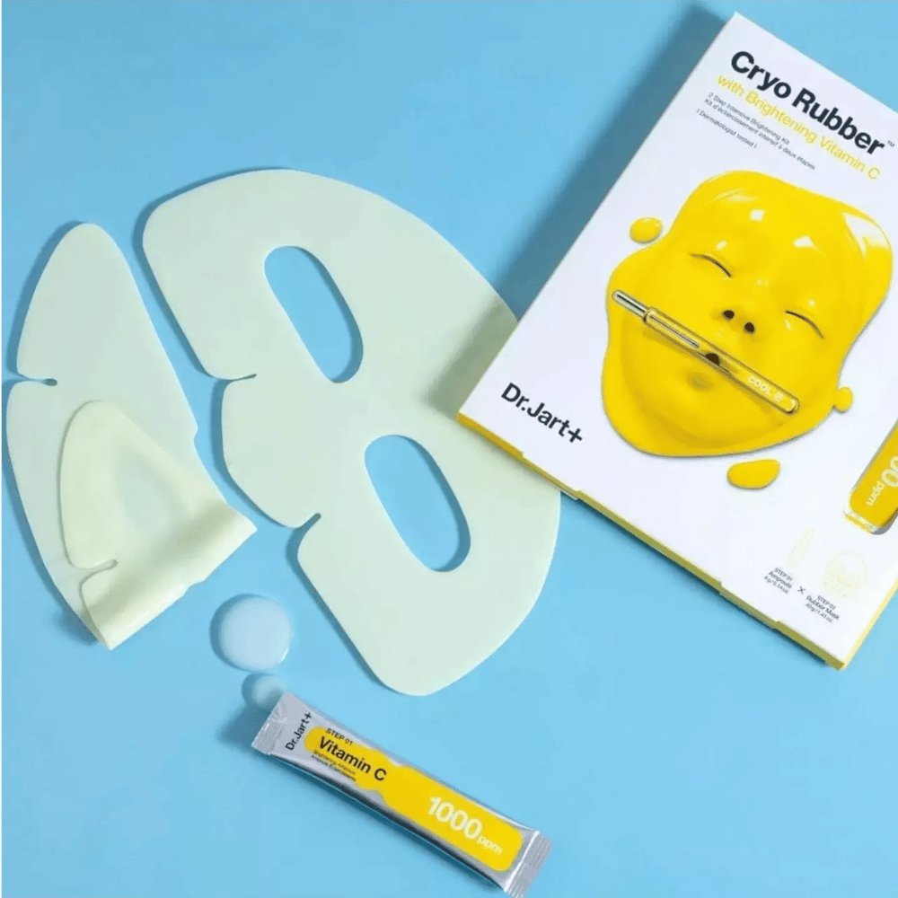 Immagine del prodotto di bellezza 'Cryo Rubber™ con Vitamina C Illuminante' di Dr.Jart+. Su uno sfondo blu si vede una maschera facciale di gomma bianca, un piccolo tubetto con la scritta 'Vitamina C 1000ppm' e una scatola con il nome del prodotto e l'immagine della maschera gialla.