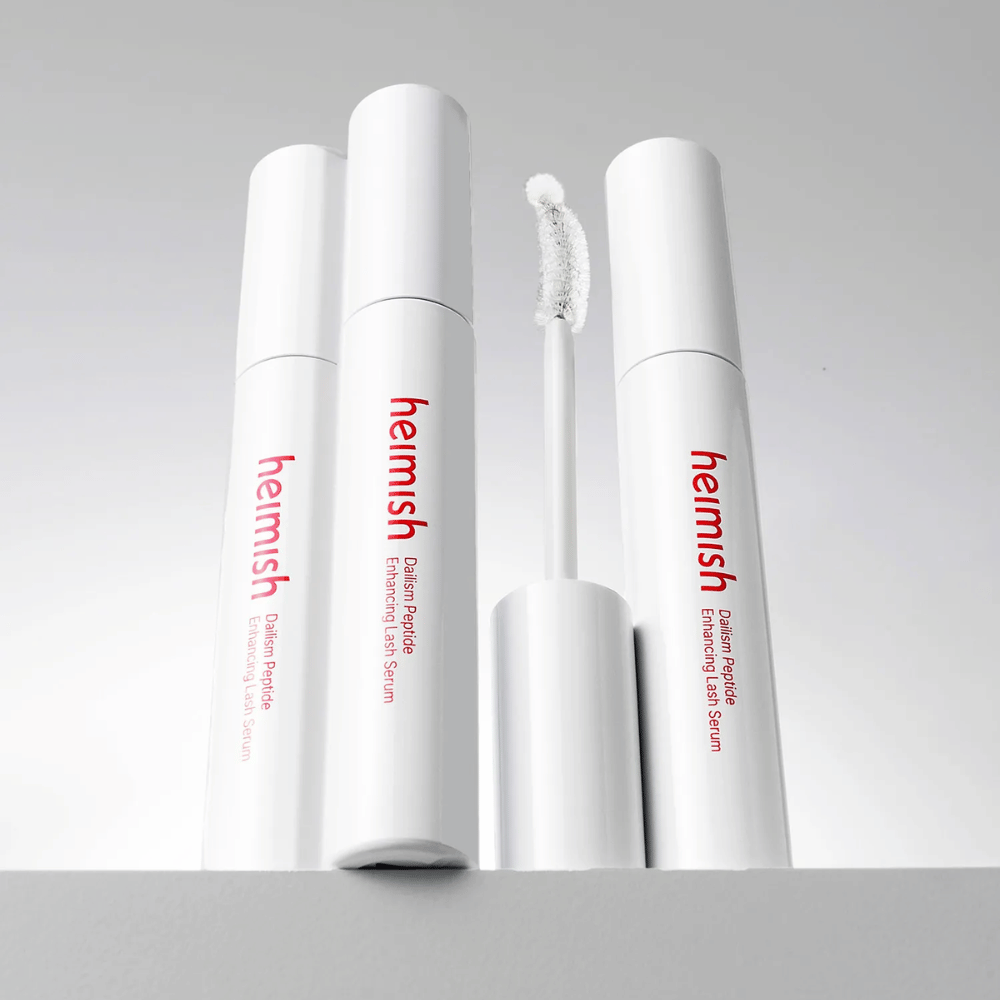 Diverse confezioni di Heimish Dailism Peptide Enhancing Lash Serum sono mostrate insieme allo scovolino applicatore curvo. L'immagine mette in evidenza il design elegante del prodotto e lo scovolino di precisione per un'applicazione facile.