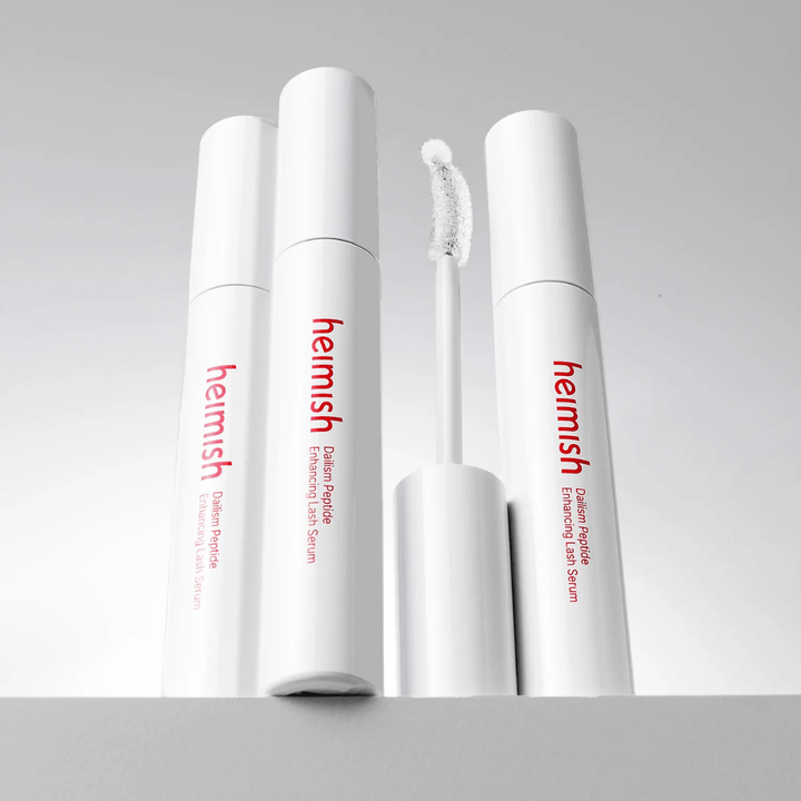 Diverse confezioni di Heimish Dailism Peptide Enhancing Lash Serum sono mostrate insieme allo scovolino applicatore curvo. L'immagine mette in evidenza il design elegante del prodotto e lo scovolino di precisione per un'applicazione facile.