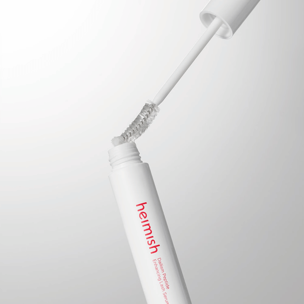 Primo piano dell'applicatore di Heimish Dailism Peptide Enhancing Lash Serum. Lo scovolino viene estratto dal contenitore bianco e mostra la formula trasparente del siero che viene applicata sulle ciglia.