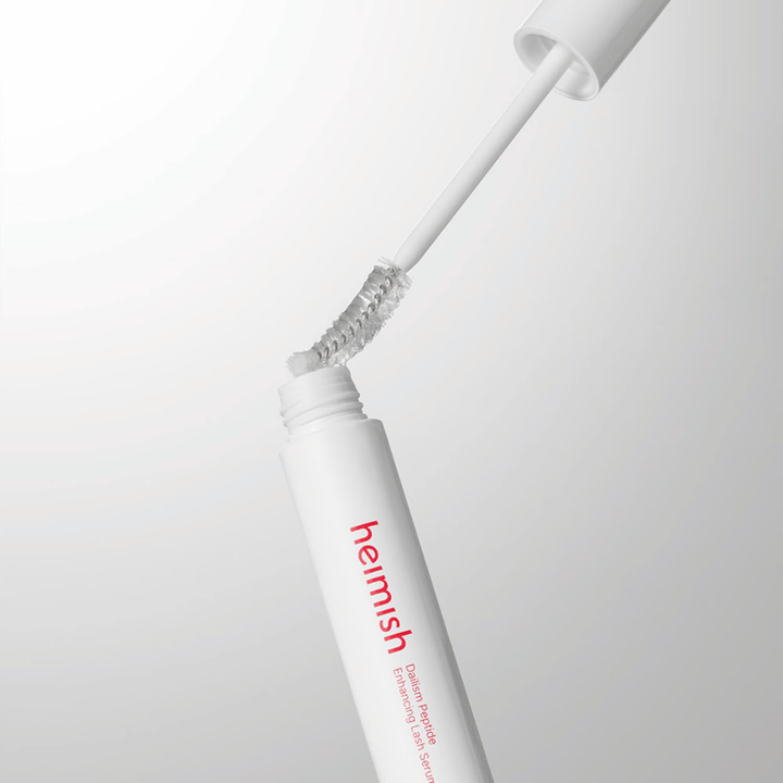 Primo piano dell'applicatore di Heimish Dailism Peptide Enhancing Lash Serum. Lo scovolino viene estratto dal contenitore bianco e mostra la formula trasparente del siero che viene applicata sulle ciglia.