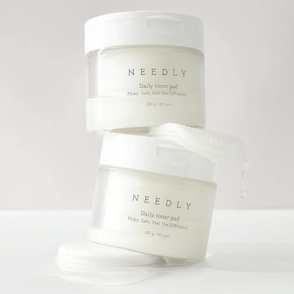 Due barattoli con Needly Daily Tonico viso Pad impilati uno sopra l'altro. I barattoli sono bianchi con testo minimalista e contengono ciascuno 60 dischetti tonificanti. Un dischetto tonificante è parzialmente fuori dal barattolo superiore con liquido che gocciola.