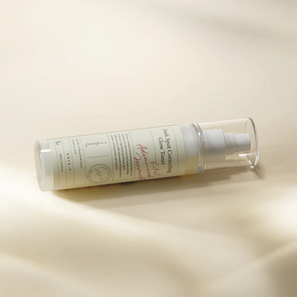 Una bottiglia distesa di "AXIS-Y Dark Spot Correcting Glow Toner" posizionata su una superficie chiara. La bottiglia è trasparente con un'etichetta bianca e ha una pompa spray bianca. L'etichetta mostra informazioni sulle proprietà schiarenti e idratanti del prodotto e contiene ingredienti chiave come Glutatione e Niacinamide.