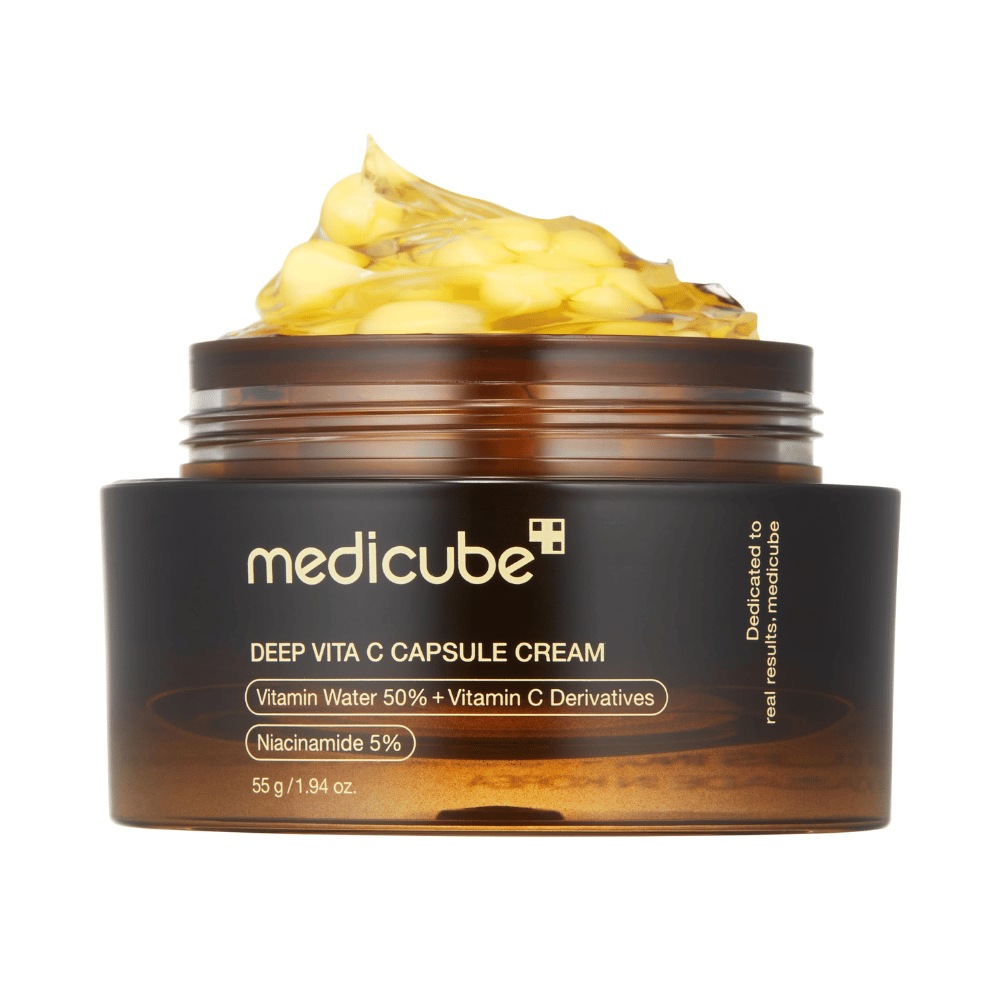 Un primo piano di "Medicube Deep Vita C Capsule Cream", che mostra la consistenza della crema e le capsule di vitamina C. La confezione è di colore marrone scuro con testo dorato e descrive il contenuto di acqua vitaminica (50%) e niacinamide (5%), offrendo una presentazione visiva delle proprietà idratanti e migliorative della pelle del prodotto.