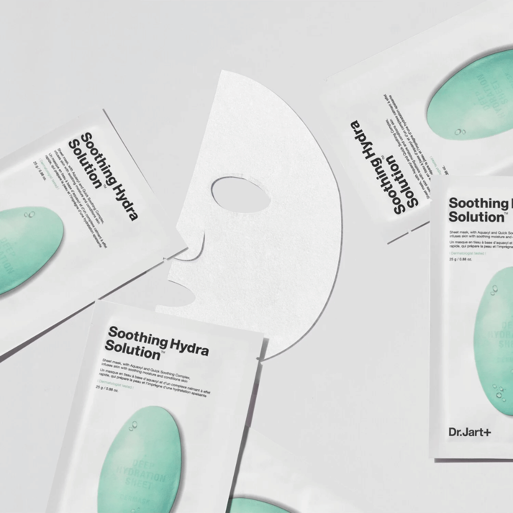 Diverse confezioni di maschere viso "Soothing Hydra Solution" da Dr.Jart+ sono sparse su una superficie chiara. Una maschera aperta è posizionata al centro, mostrando la sua struttura bianca e perforata.