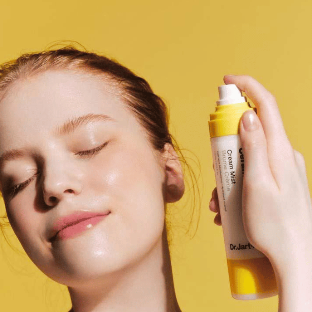 Una donna tiene una bottiglia Dr. Jart+ Ceramidin Cream Mist 110ml vicino al viso, promuovendo i suoi benefici di aumento dell'umidità e la formulazione vegana.