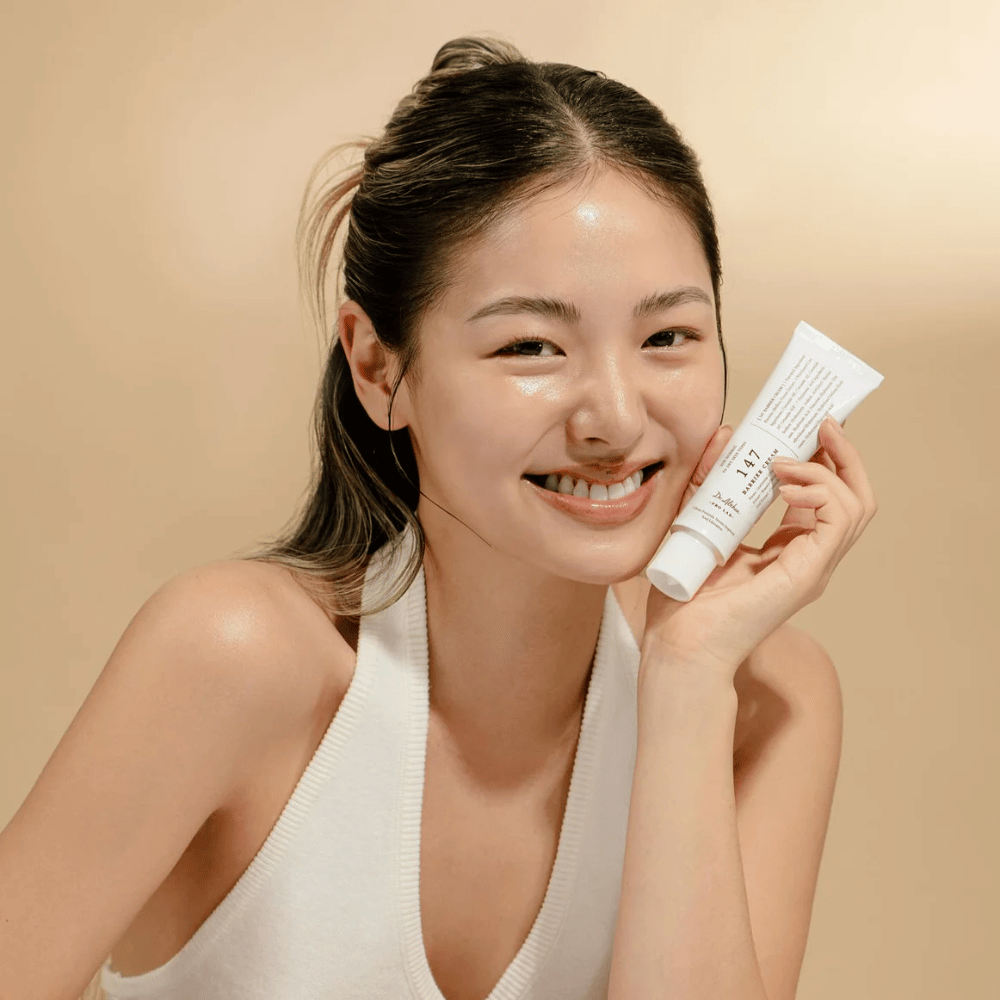 Modello con pelle luminosa che sorride e tiene in mano Dr. Althea 147 Barrier Cream – una crema viso coreana per la barriera cutanea e l'idratazione.