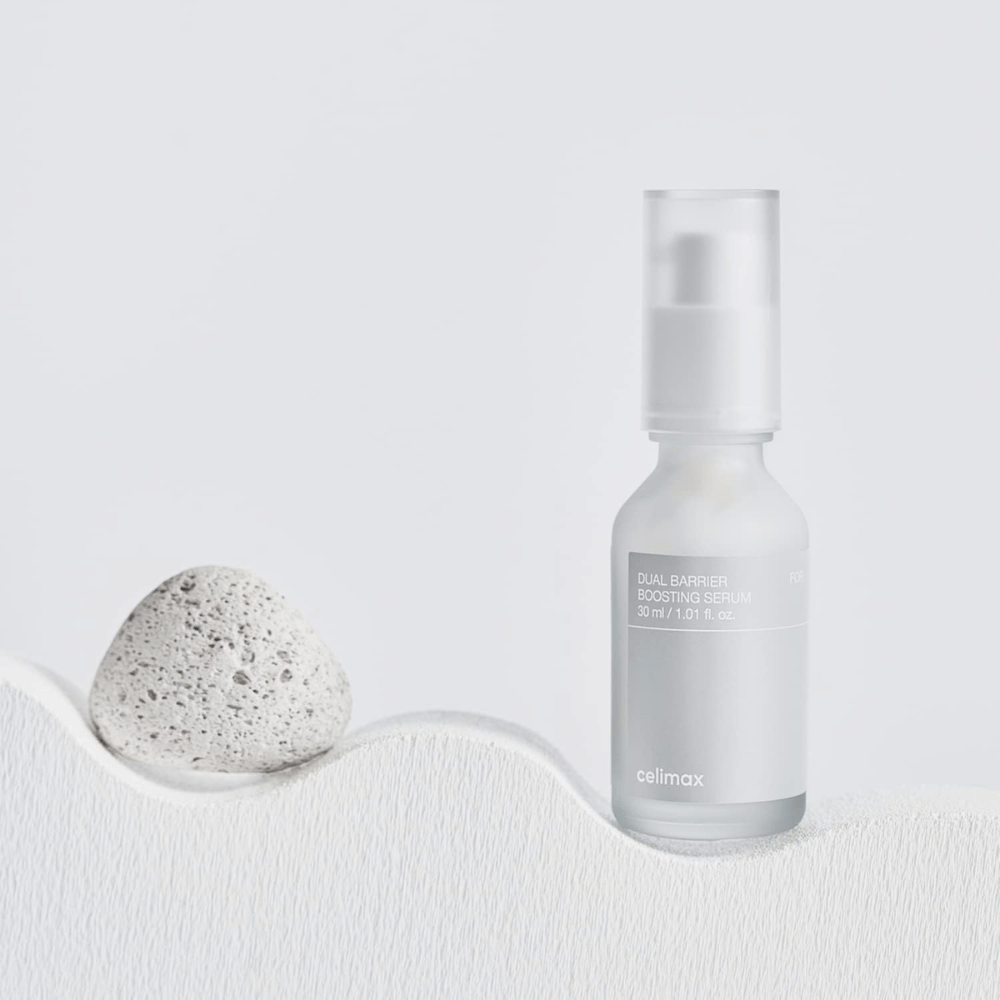 Celimax Dual Barrier Boosting Serum posizionato su tessuto con pietra, immagine del prodotto tranquilla che segnala idratazione e barriera cutanea rafforzata.