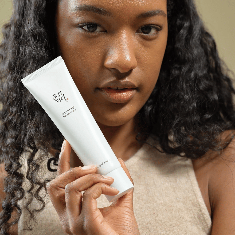 Una donna con capelli ricci tiene un tubetto di "Beauty of Joseon Dynasty Cream" vicino al viso, mettendo in risalto il prodotto. La crema, presentata in un elegante tubetto bianco, è progettata per nutrire e ringiovanire la pelle, sottolineando la sua attrattiva per diversi tipi di pelle.