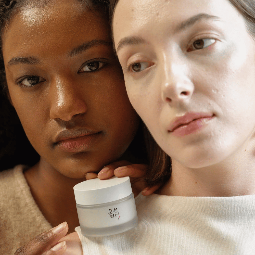 Due donne, una con carnagione più scura e una con carnagione più chiara, stanno vicine con l'attenzione sui loro volti e una crema per il viso di "Beauty of Joseon". La crema è posizionata davanti a loro, enfatizzando il suo utilizzo per diversi tipi di pelle e la sua capacità di donare luminosità e nutrimento.