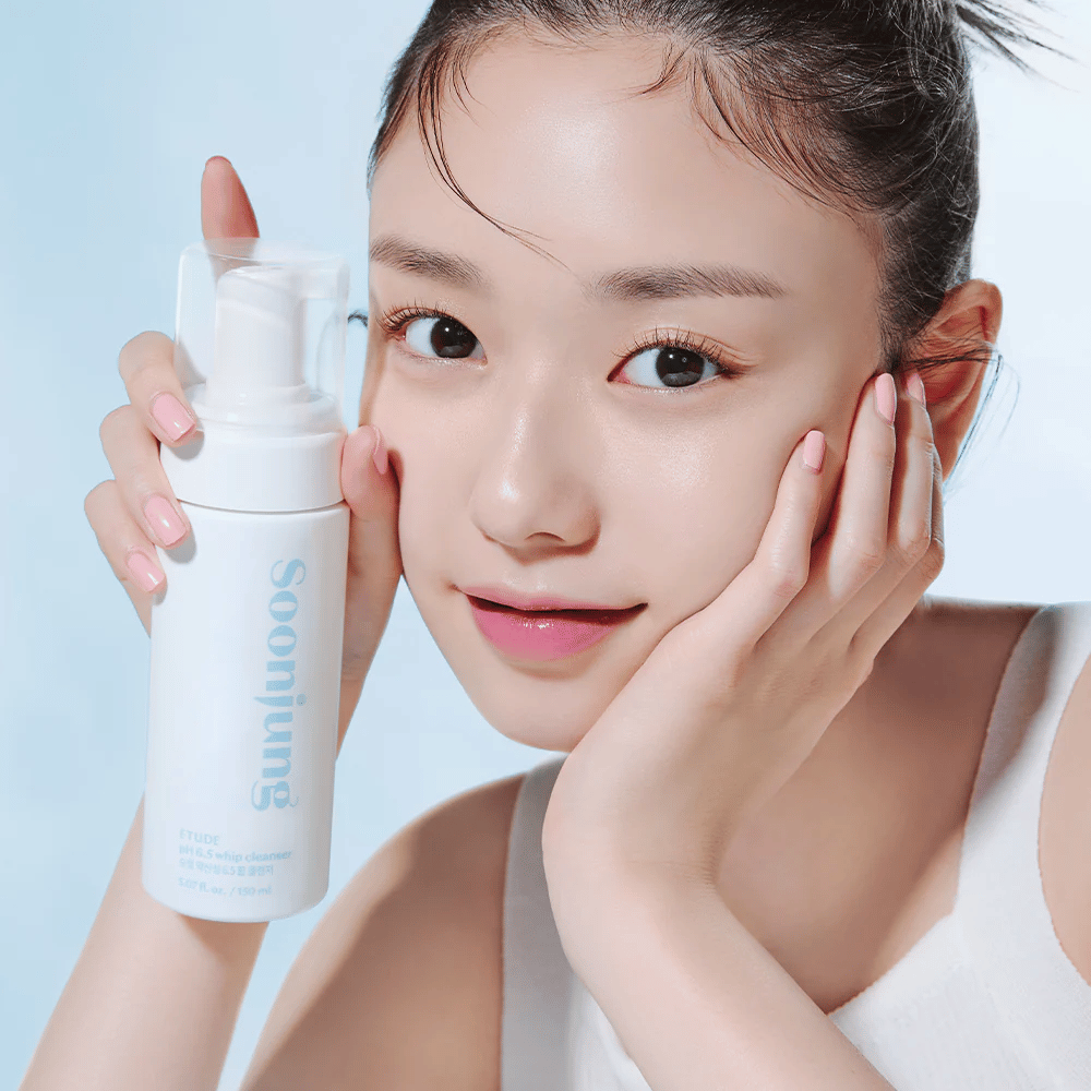 Una giovane donna con una pelle chiara tiene un flacone bianco con pompa con il testo "Soon Jung pH 6.5 Whip Cleanser" da ETUDE. Sorride alla fotocamera con un leggero trucco e un aspetto fresco su uno sfondo azzurro.