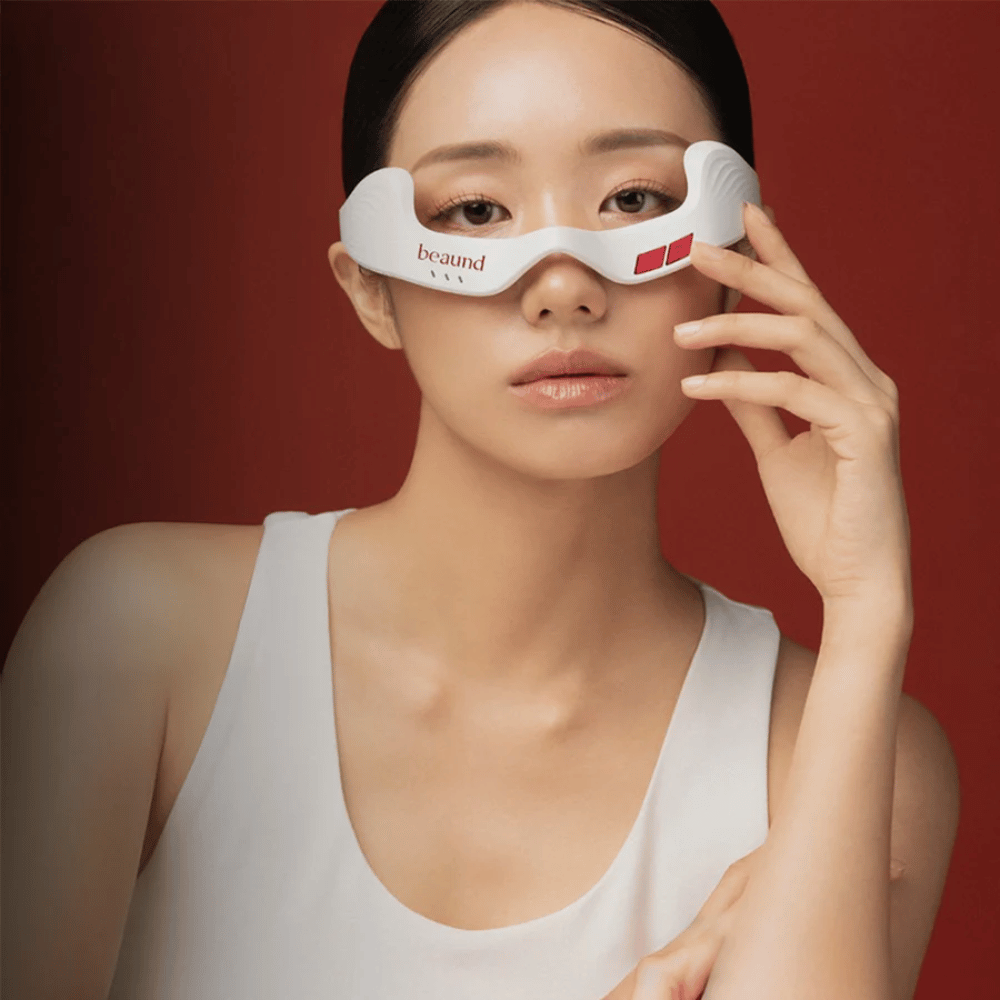 Immagine con modella in cui una persona utilizza Beaund Eye Rejuv, un dispositivo LED futuristico per l’area intorno agli occhi, su un elegante sfondo rosso.