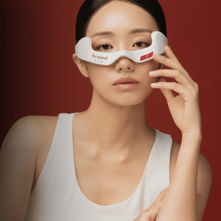 Immagine con modella in cui una persona utilizza Beaund Eye Rejuv, un dispositivo LED futuristico per l’area intorno agli occhi, su un elegante sfondo rosso.