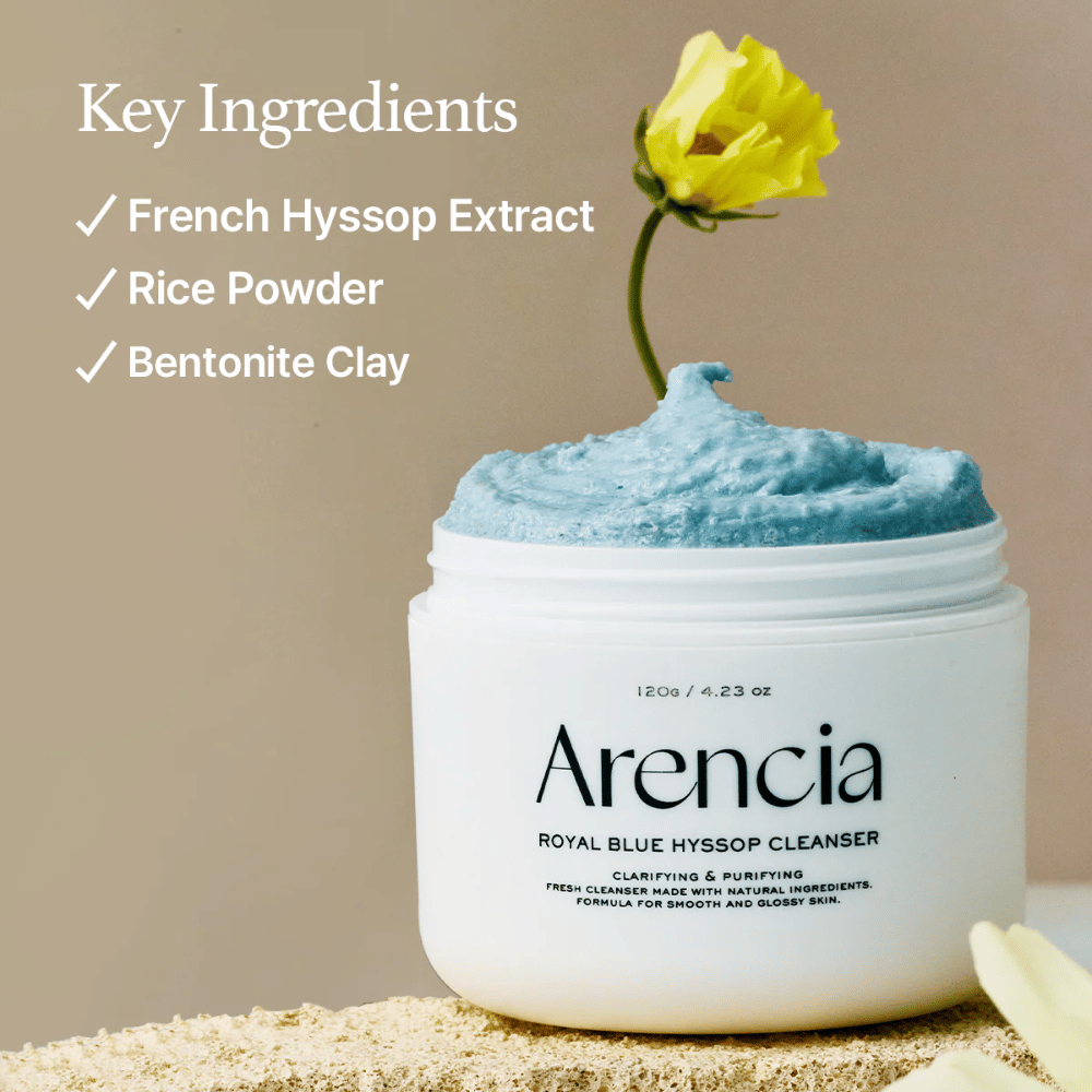 Immagine del barattolo "Arencia Royal Blue Hyssop Cleanser" riempito con crema detergente blu. Un fiore giallo spunta dalla crema. Il testo nell'immagine evidenzia tre ingredienti chiave: Estratto di Issopo Francese, Polvere di Riso e Argilla di Bentonit.