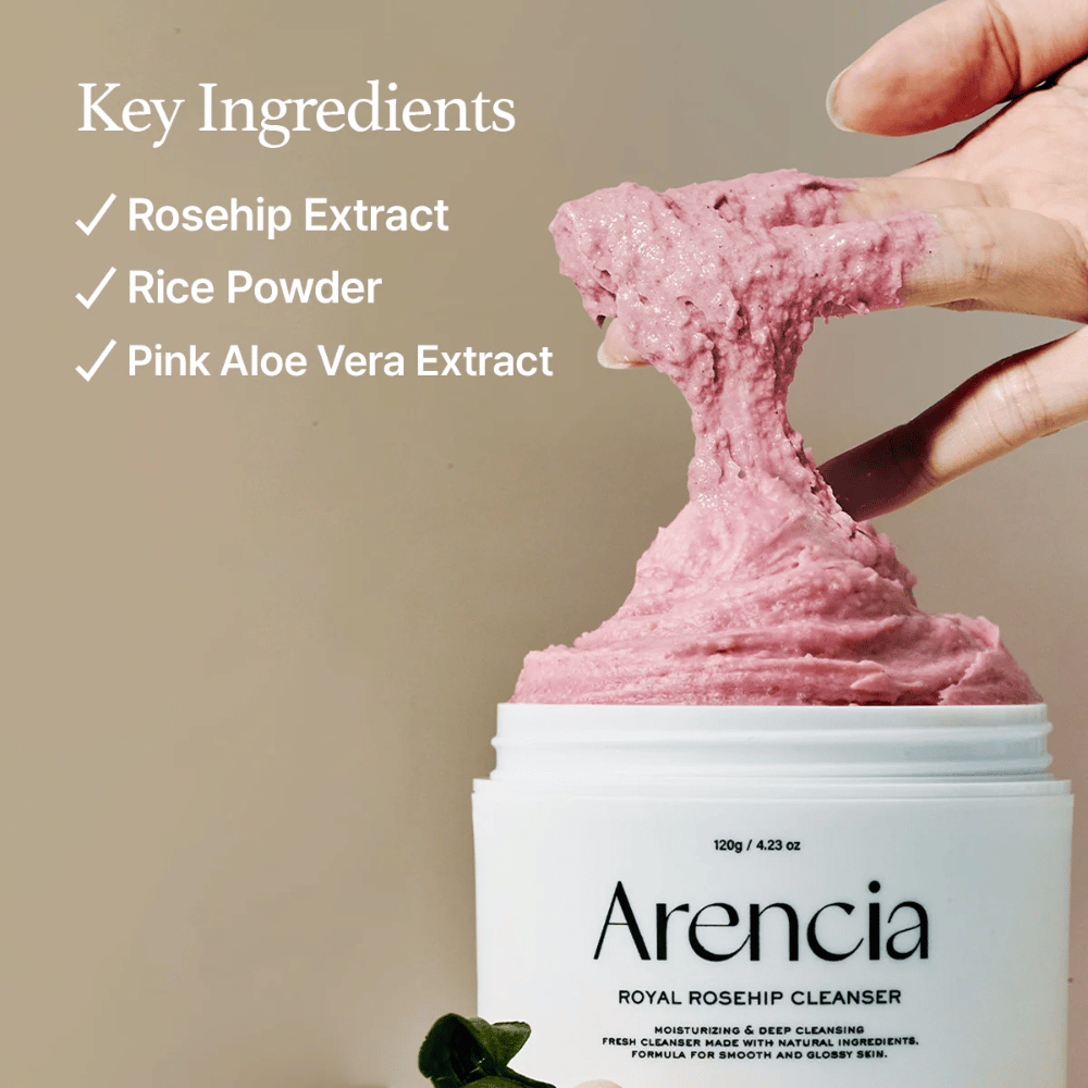 Una mano solleva la texture detergente rosa e soffice dal barattolo Arencia Royal Rosehip Cleanser, con la lista degli ingredienti a sinistra: estratto di rosa canina, polvere di riso ed estratto di aloe vera rosa.