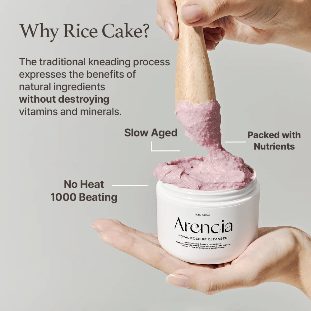 Una mano tiene un barattolo aperto di Arencia Royal Rosehip Cleanser, con una formula detergente rosa simile a mochi sollevata con una spatola di legno – mostra la texture del prodotto e il metodo di produzione tradizionale.