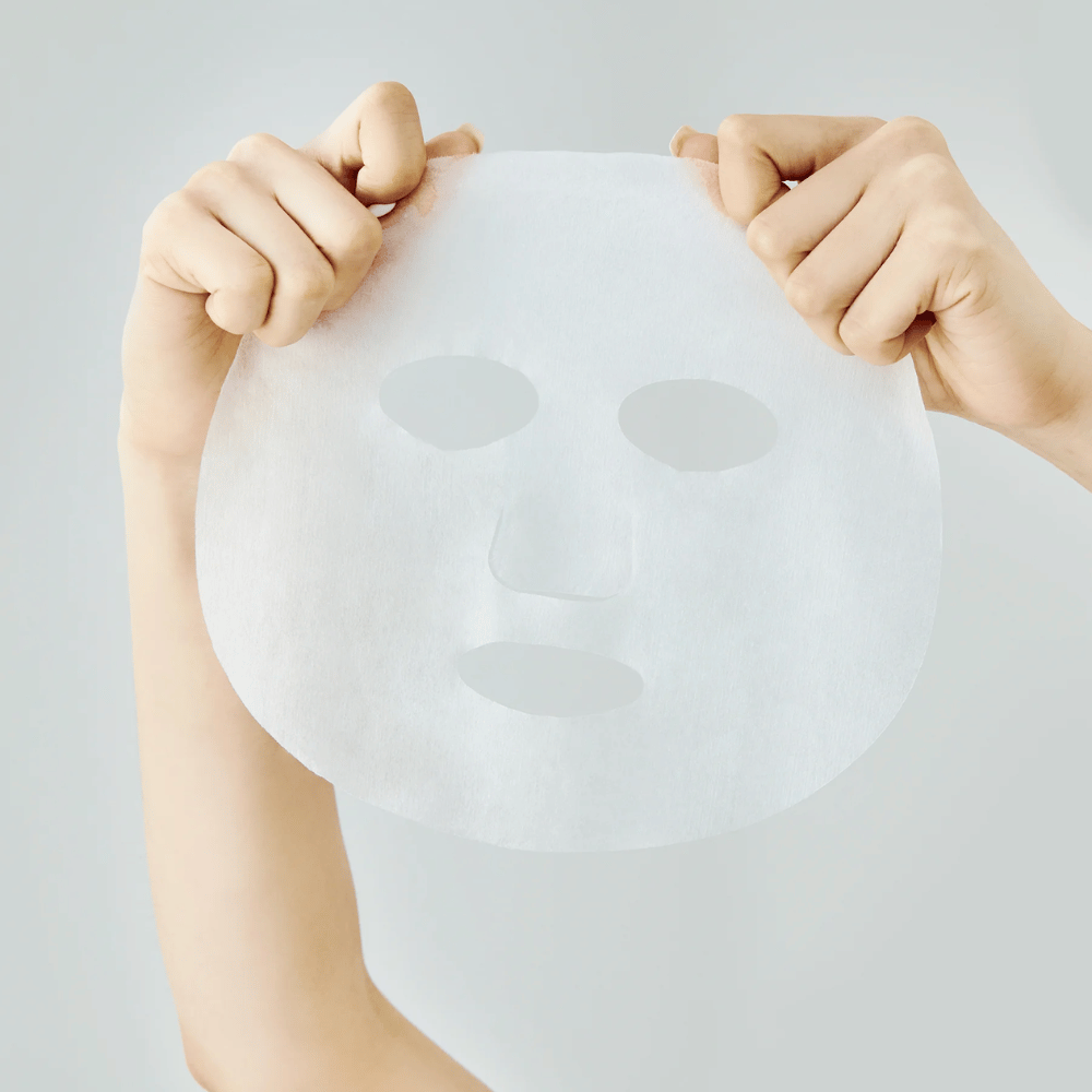 Due mani tengono una maschera in tessuto bianca su uno sfondo grigio chiaro, con ritagli visibili per occhi, naso e bocca.