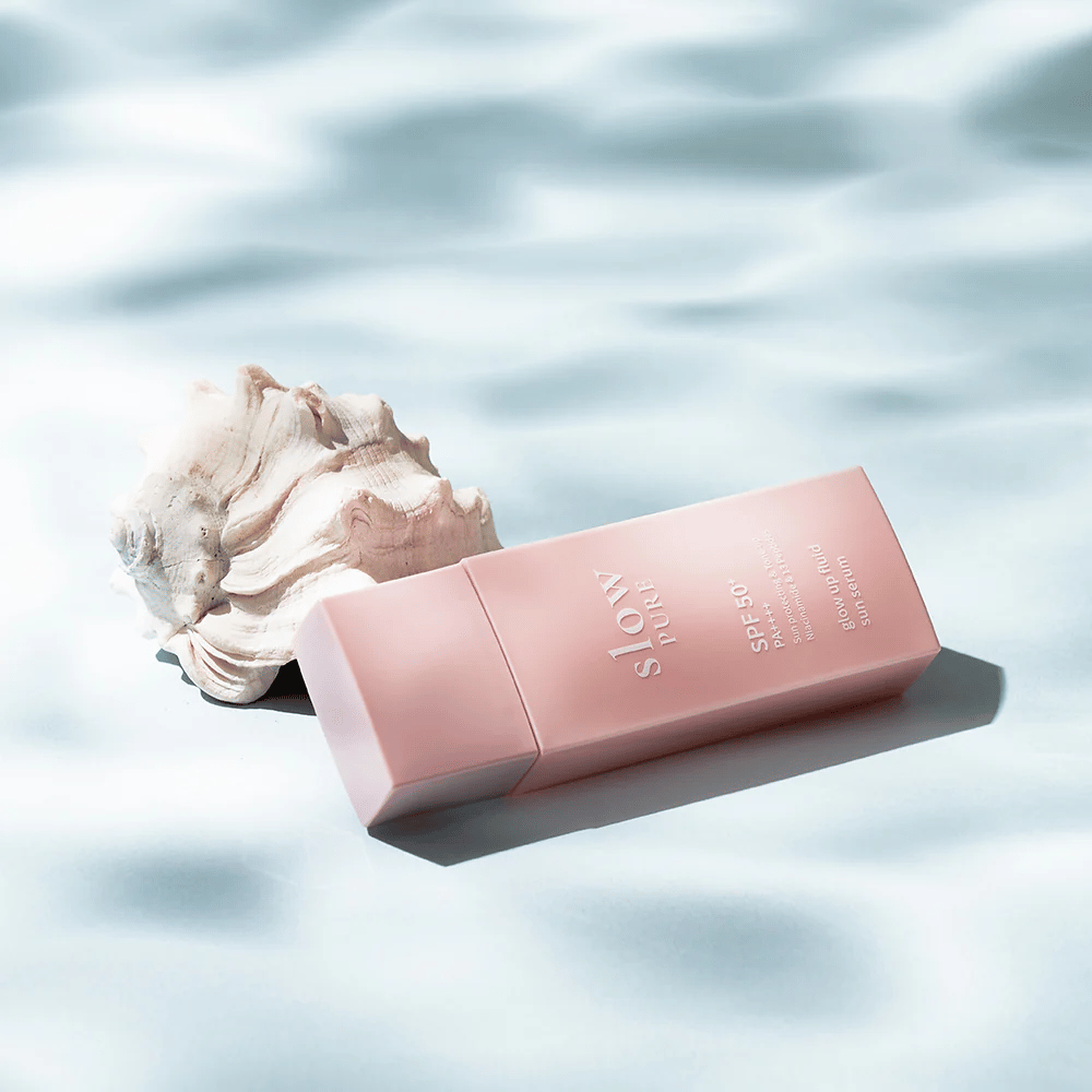 Slow Pure Glow Up Fluid Sun Serum vicino a conchiglie e effetto acqua, evidenzia protezione solare e luminosità.