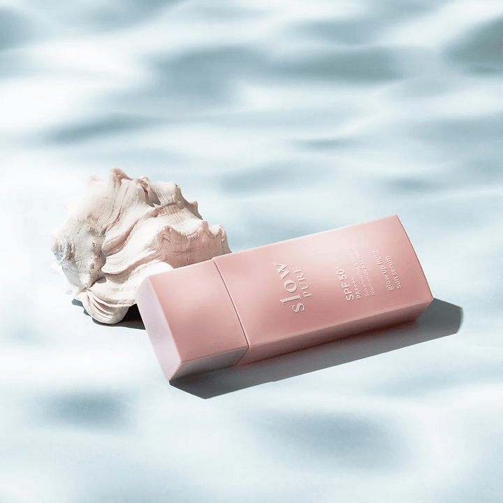 Slow Pure Glow Up Fluid Sun Serum vid snäckskal och vatteneffekt, framhäver solskydd och lyster.
