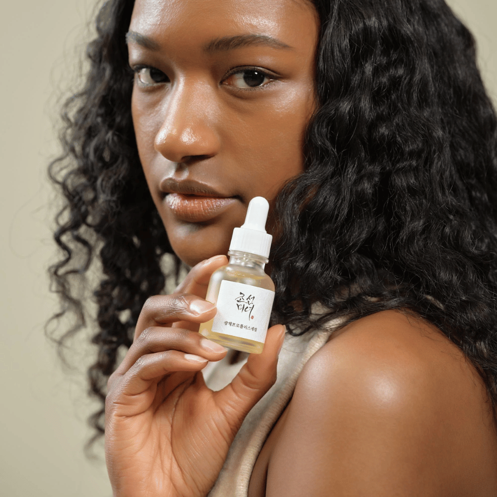 Donna con capelli ricci che solleva un flacone di Beauty of Joseon Glow Serum con pipetta, un prodotto per la cura della pelle idratante e illuminante.