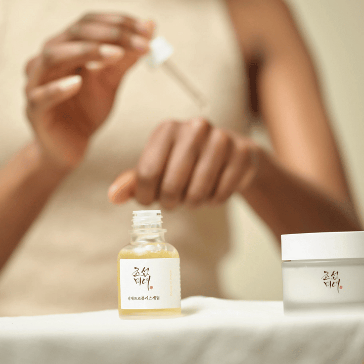 Person som applicerar Beauty of Joseon Glow Serum med pipett från glasflaska, med en burk av samma serie bredvid på bordet.