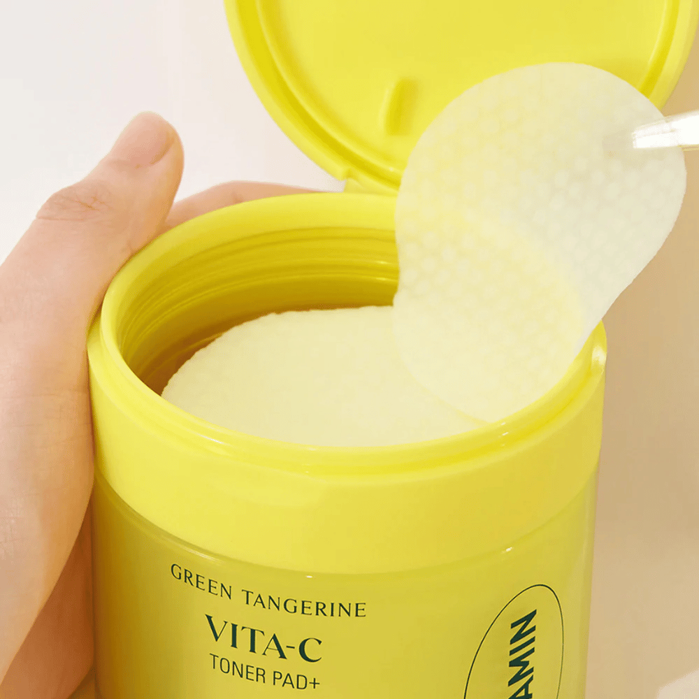 Una mano utilizza una pinzetta di plastica per prendere un dischetto tonico con texture da un contenitore giallo chiaro etichettato "GREEN TANGERINE VITA-C TONER PAD+" del marchio "gooda