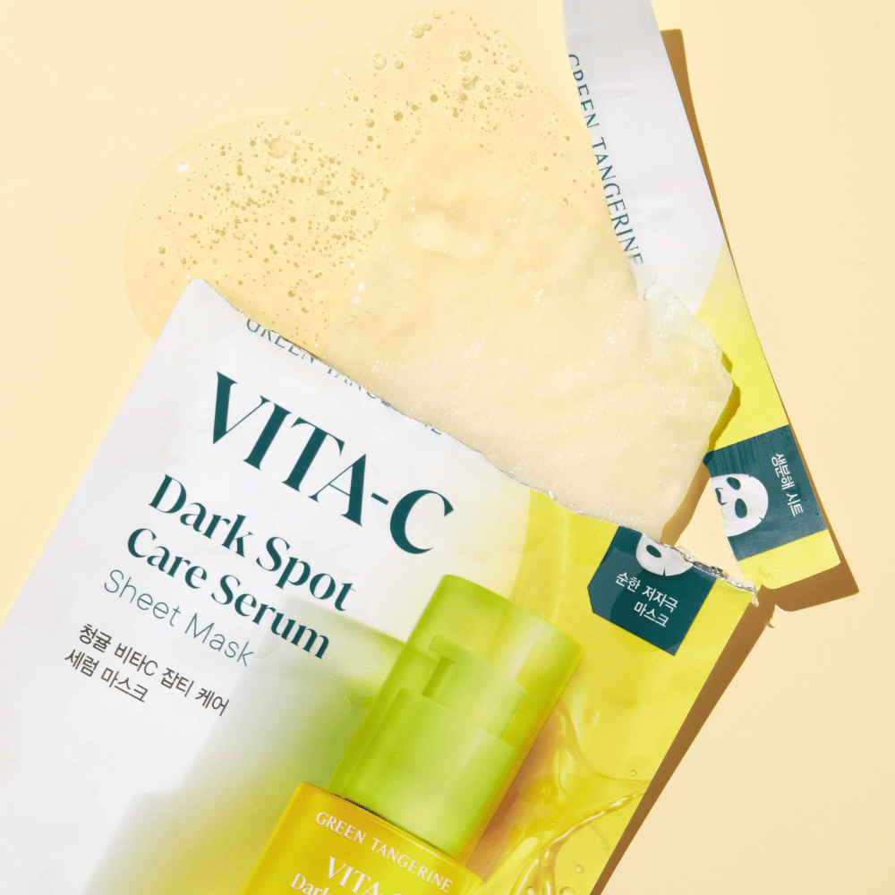 Confezione aperta della maschera in foglio VITA-C Dark Spot Care Serum con una maschera viso sopra e una bottiglia di siero verde, il tutto su uno sfondo giallo.