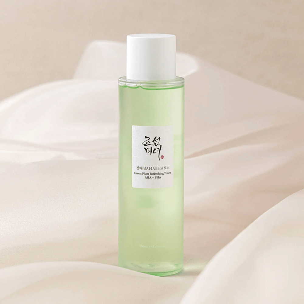 Una bottiglia di "Green Plum Refreshing Tonico viso" AHA + BHA si trova su uno sfondo morbido e chiaro. Il prodotto ha un liquido verde che riflette freschezza e naturalezza. L'etichetta è bianca con dettagli neri e rossi, che conferiscono un aspetto elegante e pulito.