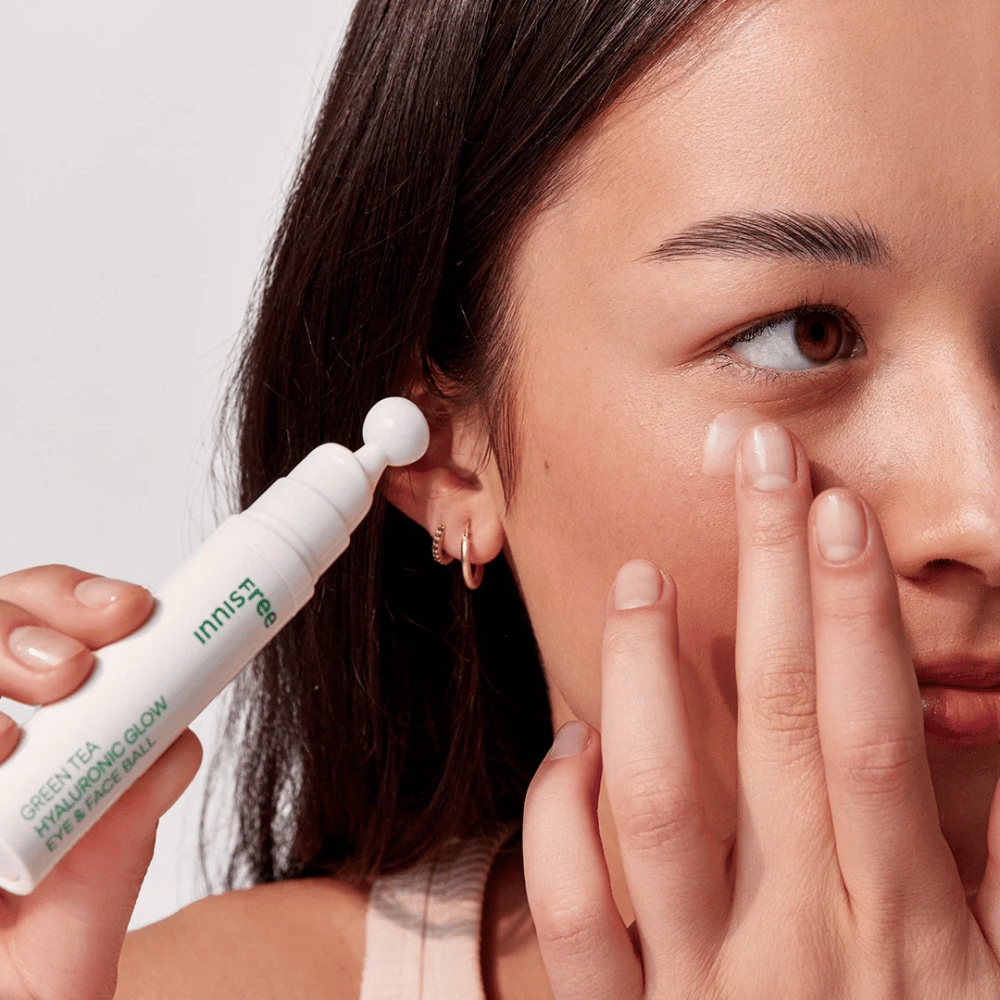 Una persona applica Innisfree Green Tea Hyaluronic Glow Eye & Face Ball sulla pelle sotto gli occhi. Il prodotto ha una parte applicatrice bianca e rotonda ed è in una bottiglia bianca con testo verde. Il viso dell'utente è parzialmente visibile e si concentra sull'applicazione del prodotto.