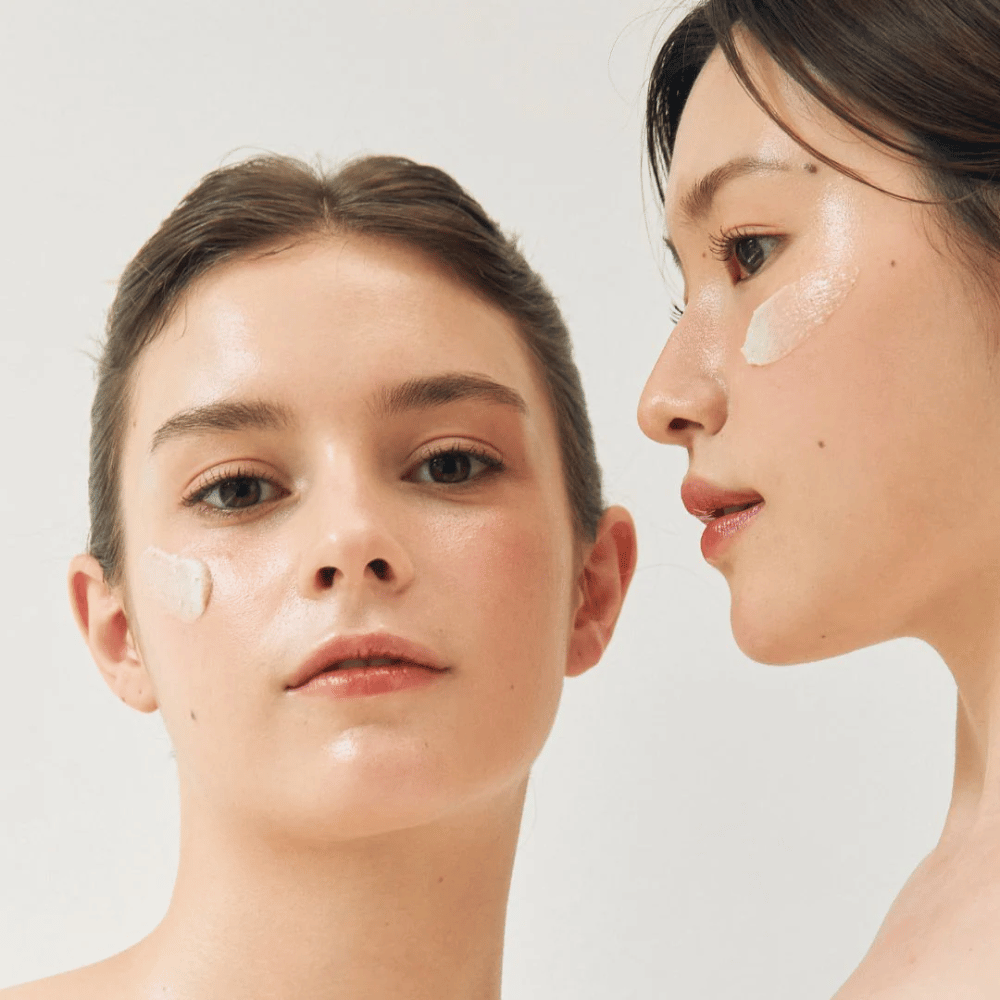 Due donne con maschere facciali applicate su parti dei loro volti. Una donna ha la maschera sulla guancia e sul naso, mentre l'altra ce l'ha sotto l'occhio e sulla guancia. Hanno un trucco naturale e sembrano fresche, il che mette in risalto l'effetto idratante e illuminante delle maschere.