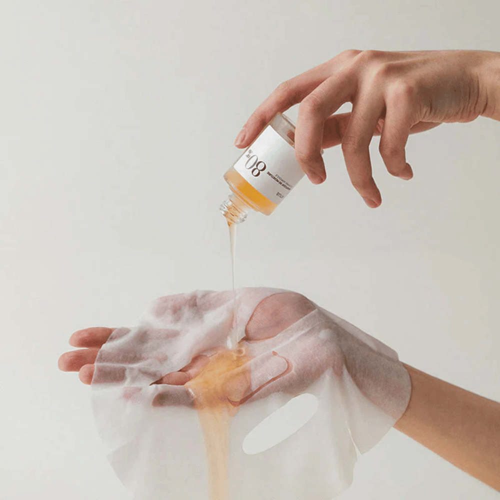 Una mano versa un liquido dorato e denso da una piccola bottiglia con l'etichetta '108' su un maschera viso trasparente tenuto in un'altra mano. Lo sfondo è neutro e uniforme per mettere in risalto il prodotto.