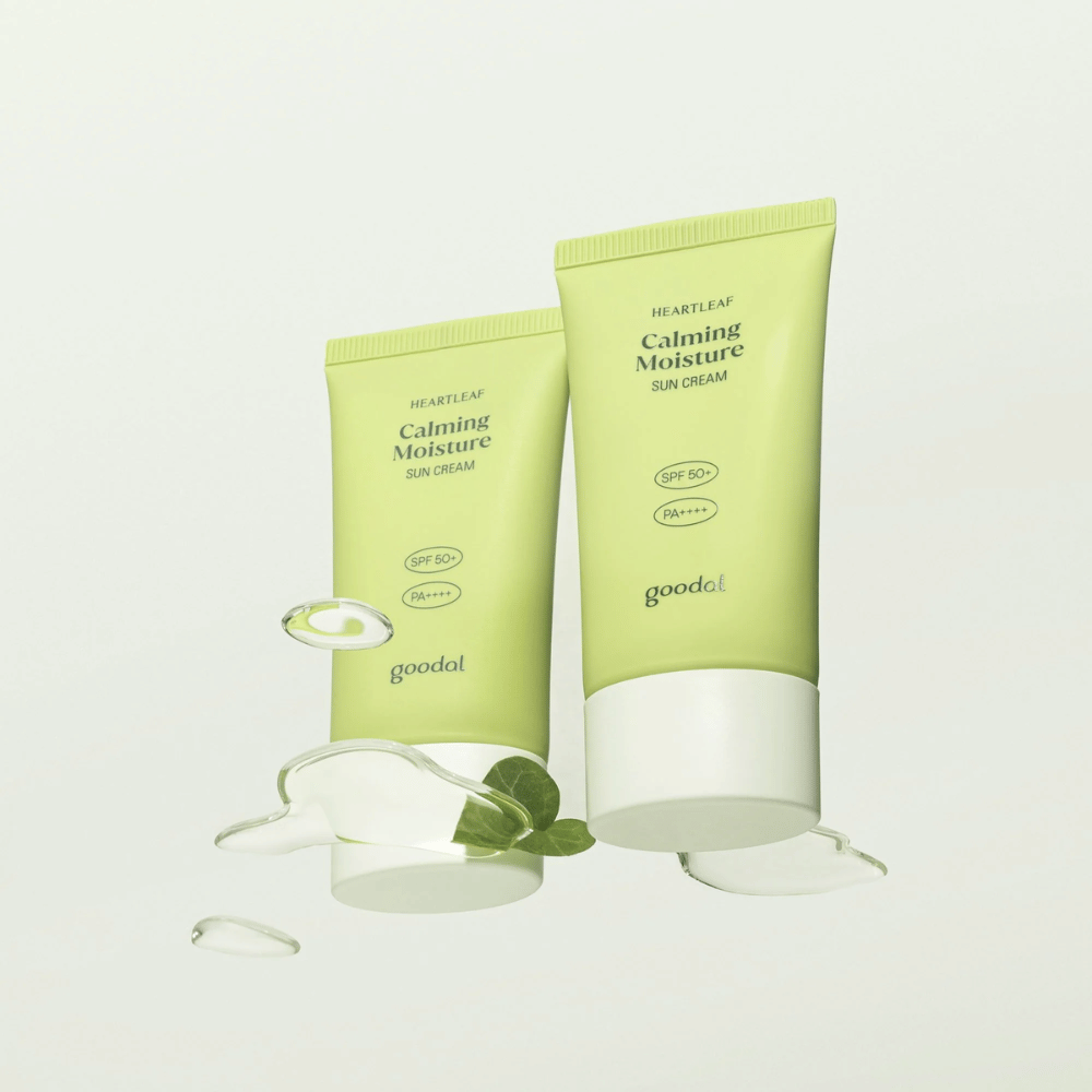 Due tubi di Goodal Heartleaf Calming Moisture Sun Cream SPF50+ PA++++ circondati da foglie di piante e gocce di siero su sfondo chiaro.