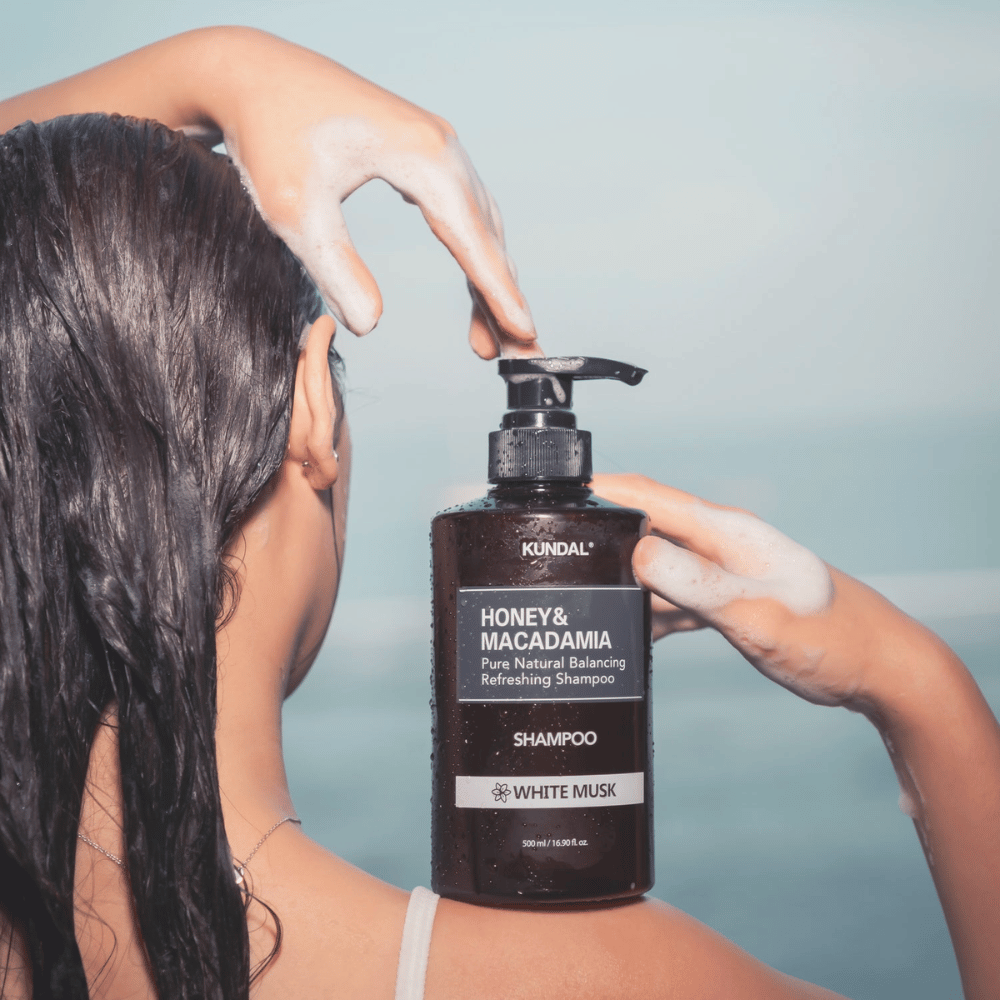 Donna con capelli bagnati applica schiuma e tiene KUNDAL Honey & Macadamia Nature Shampoo White Musk con sfondo del mare.